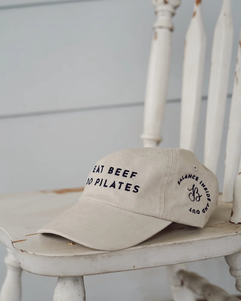 The cream Balance Inside and Out cap, embroidered “Eat beef, do Pilates”, resting on a white chair