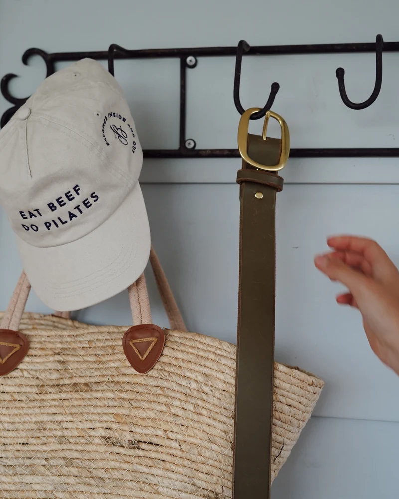 The cream “Eat beef, do Pilates” cap hanging on a hook beside a woven basket and a leather belt