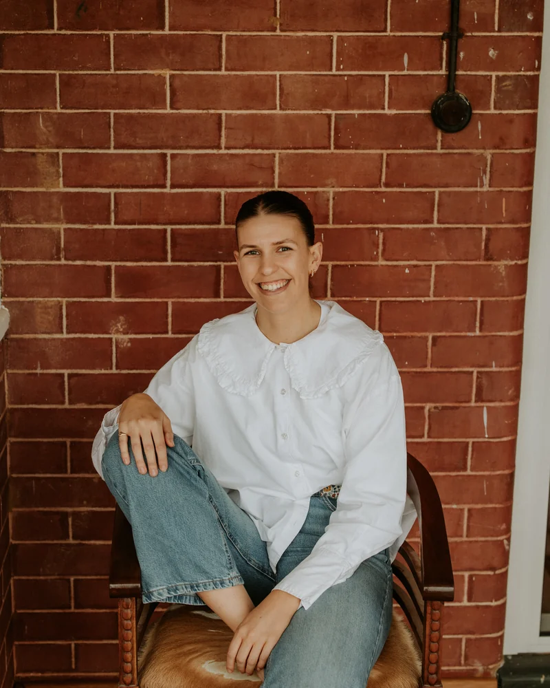 Laura Kiel smiling, sitting on a timber chair against a red brick wall