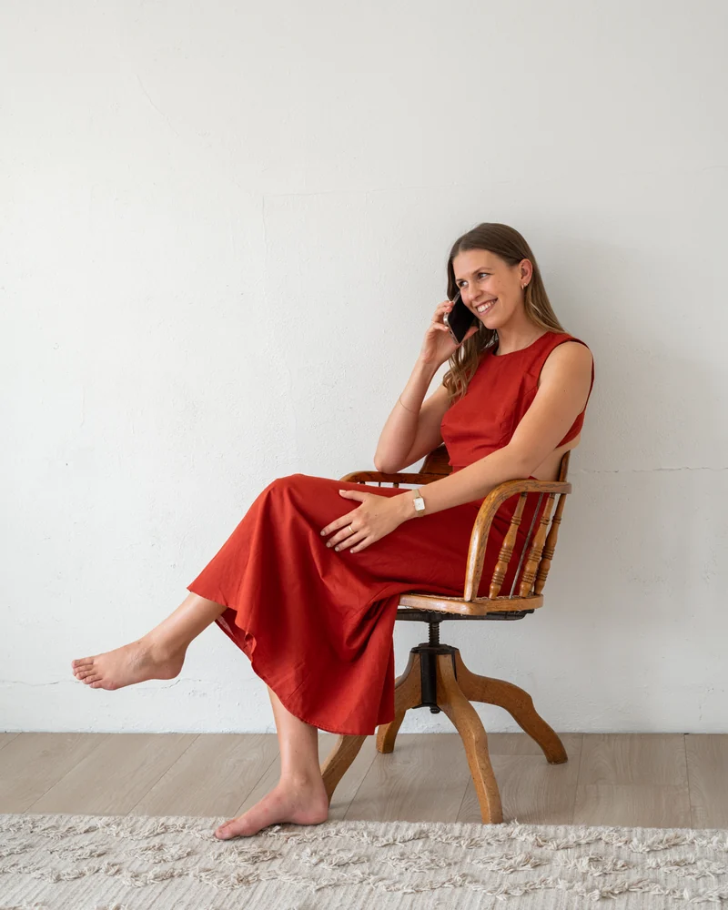 Laura laughing on the phone, sitting on a timber chair in a red dress