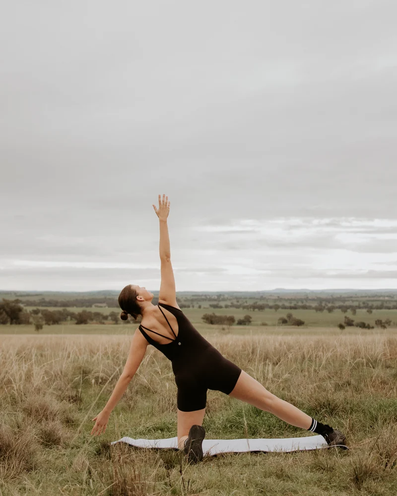 Laura holding a side-stretch Pilates pose on a mat in an open paddock
